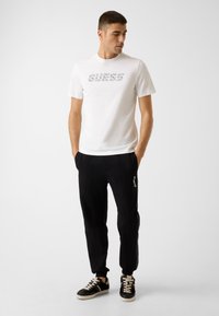 Weißes T-Shirt mit kurzen Ärmeln und "GUESS"-Logo, kombiniert mit schwarzen, schmal zulaufenden Hosen mit weißen Seitenstreifen und schwarzen Sportschuhen.