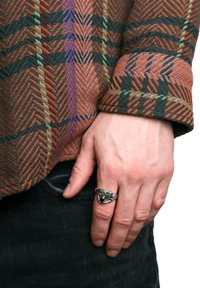 Chemise tissée multicolore avec un motif en zigzag dans des tons de vert, orange et marron. Bague en argent avec une pierre sombre sur une main reposant sur un jean noir.