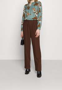 Blouse teal à motif floral avec un détail en forme de clé, associée à un pantalon marron taille haute. Le mannequin porte des bottes à talons noirs et tient un petit sac noir.