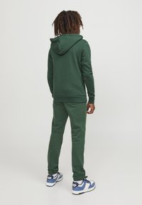 Grön hoodie och matchande joggers i mjukt tyg, med huva, ribbade ärmslut och sömdetaljer, kombinerat med vita och blå sneakers.