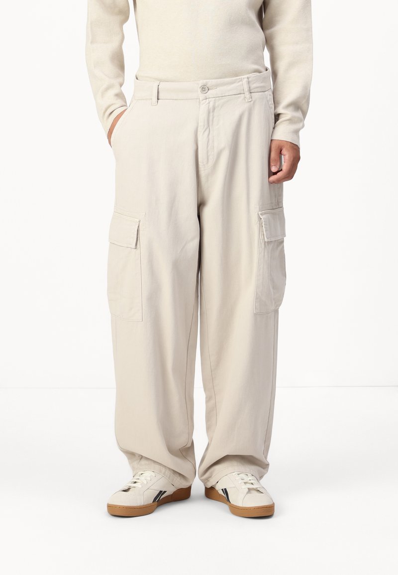 Pantaloni cargo bej, cu o croială lejeră, două buzunare laterale și închidere cu nasture, asortați cu adidași albi și un pulover deschis la culoare.