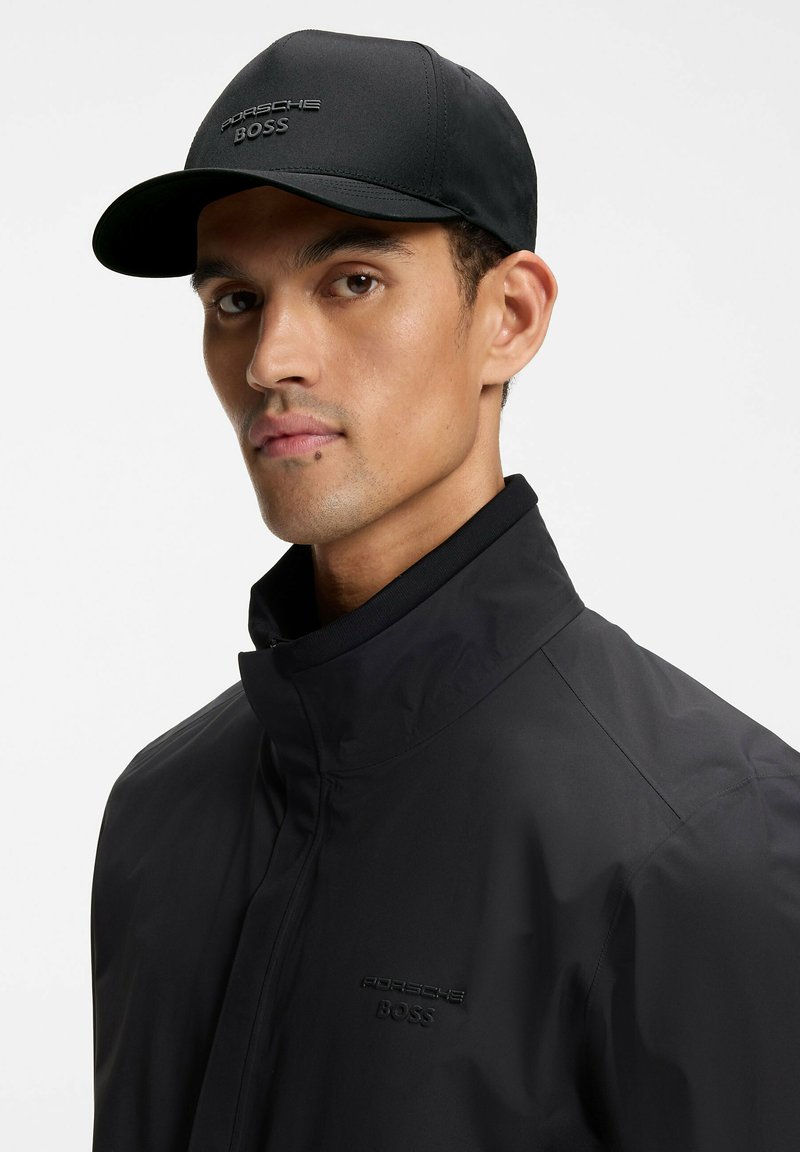 Man die een zwarte Porsche BOSS-pet en bijpassende zwarte jas met hoge kraag draagt, kijkt iets naar de zijkant tegen een effen achtergrond.