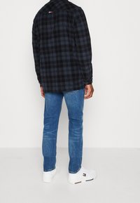 Uomo vestito con giacca a quadri nera e blu, jeans blu e sneakers bianche, in piedi contro un muro bianco, rivolto di spalle verso la macchina fotografica.