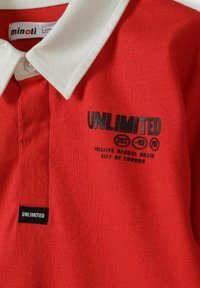 Czerwony podkoszulek polo z białym kołnierzykiem, z czarnym nadrukowanym napisem "UNLIMITED" i dodatkowymi detalami. Wykonany z fakturowanej bawełny.