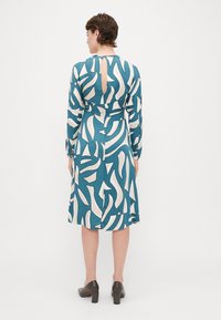 Tyrkysové šaty s bielym abstraktným vzorom, dlhé rukávy a detail kľúčovej diery na chrbte. Hladká látka s lichotivým A-liniovým strihom.