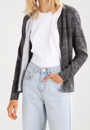 Cardigan grigio testurizzato con apertura frontale, indossato sopra una T-shirt bianca. Abbinato a jeans azzurri a vita alta. Tessuto morbido e vestibilità rilassata.