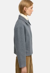 Szary, wełniany żakiet w mieszance tkanin z kołnierzykowym dekoltem, opadającymi ramionami i kieszeniami bocznymi. Projekt charakteryzuje się skróconą długością i miękką fakturą.