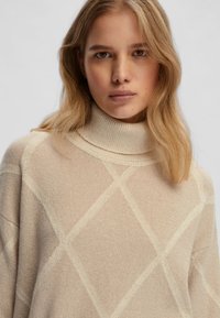 Maglione a collo alto beige chiaro con un motivo a rombi, dotato di un colletto a coste e di un materiale in maglia morbido e testurizzato.