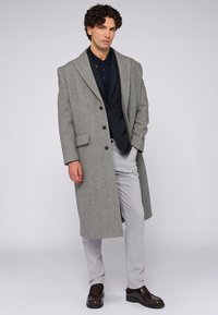 Šedý vlněný kabát s klopovými límci, třemi knoflíky a postranními kapsami, kombinovaný s tmavou košilí a světle šedými kalhotami.