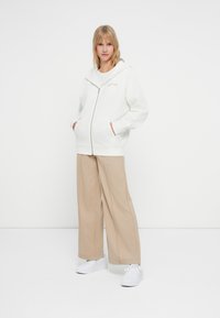 Sudadera blanca con cremallera y un corte oversized, con bolsillos y ribetes en contraste, combinada con pantalones beige de pierna ancha y zapatillas blancas.