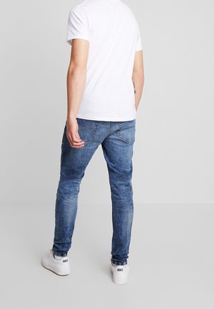 Mand iført hvid t-shirt med korte ærmer, smalle blå jeans og hvide ASICS sneakers, stående mod en almindelig lysegrå baggrund.