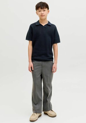 Mladić stoji noseći crnu teksturiranu polo majicu kratkih rukava, sive hlače i bež casual cipele na svijetloj pozadini.