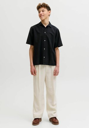 Mladý muž v čiernej krátkorukávovej košeli na gombíky, vo voľných bielych nohaviciach a hnedých topánkach, stojaci pred jednoduchým svetlým pozadím.