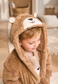 Ein kleines Kind trägt einen weichen, braunen Bärenkapuzenmantel, lächelt und schaut leicht nach unten in einer gemütlichen Innenumgebung.