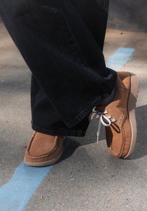 Persona con zapatos de ante marrón y pantalones negros de pie sobre la acera con una marca de línea azul visible en el suelo.