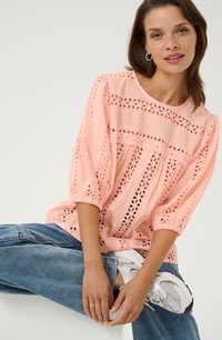 Blusa de mezcla en color rosa con detalles de encaje bordado, mangas abullonadas y un corte relajado, combinada con unos vaqueros de mezclilla azul claro y zapatillas blancas.