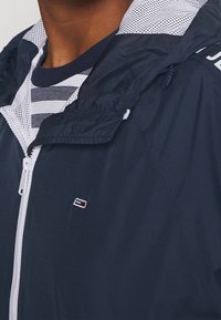 Close-up of a person wearing a navy windbreaker jacket with white zipper and small Tommy Hilfiger logo, over a navy and white striped shirt.