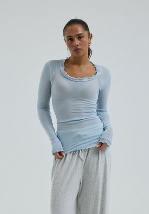 Žena oblečená v svetlomodrom tričku s dlhými rukávmi s volánmi na výstrihu a manžetách, v kombinácii s voľnými svetlosivými nohavicami, stojaca pred jednoduchým pozadím.