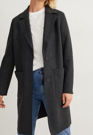 Femme portant un long manteau gris foncé sur une chemise blanche et un jean bleu, debout devant un fond clair uni.