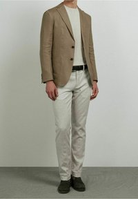 Blazer in lino marrone chiaro con rever a lancia, bottoni neri, due tasche frontali; abbinato a pantaloni grigio chiaro con trama e scarpe scure.