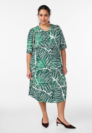 Kvinde stående i midi-kjole med grønne og hvide blade og albuelange ærmer, kombineret med mørke spidse slingback-hæle.