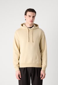 Felpa beige con texture morbida, cappuccio con cordino e un piccolo logo sul petto. Presenta polsini e orlo a costine. Abbina a pantaloni neri.