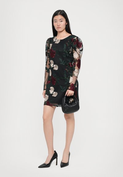 Zwarte bloemenjurk met doorschijnende lange mouwen, versierd met meerkleurige geborduurde bloemen. Model houdt een zwarte handtas vast en draagt zwarte puntige hakken.