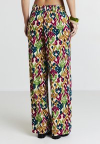 Personne portant un pantalon large multicolore avec des motifs abstraits, visible de la taille aux pieds, se tenant devant un fond uni.