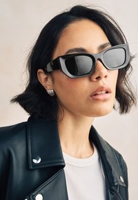 Schwarze Sonnenbrille mit dickem Rahmen, großen Gläsern und glänzendem Finish, kombiniert mit einer schwarzen Lederjacke und silbernen kreisförmigen Ohrringen.