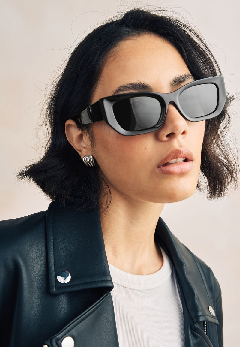 Schwarze Sonnenbrille mit dickem Rahmen, großen Gläsern und glänzendem Finish, kombiniert mit einer schwarzen Lederjacke und silbernen kreisförmigen Ohrringen.