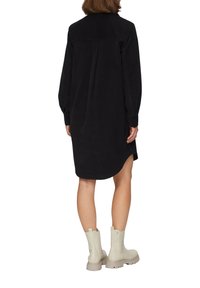 Schwarzes Cordhemdkleid mit langen Ärmeln, runder Saum und Rückenpasse, kombiniert mit cremefarbenen Knöchelschuhen. Glatte Textur, minimales Design.