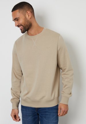 Beige sweatshirt with a crew neck, ribbed cuffs, and hem. Features textured fabric and a subtle logo on the chest. Worn with blue jeans.