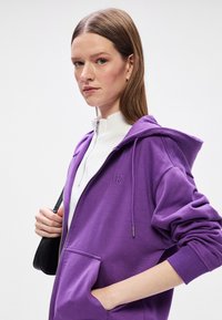 Fialová zipová mikina vyrobená z bavlny, ktorá má kapucňu so šnúrkami, predné vrecká a malé vyšívané logo na hrudi.