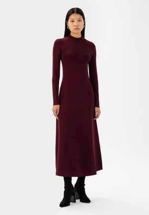 Bordeaux langemouwen tricot jurk met een aansluitend lijfje en een uitlopende rok. Model draagt zwarte enkellaarsjes met hak. Glad textuur, minimalistisch ontwerp.