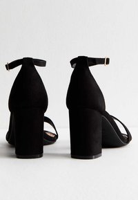Zapatos de ante negro con tacones de bloque y correas en el tobillo, que presentan una textura suave y un diseño minimalista. Las correas se cruzan sobre el pie.