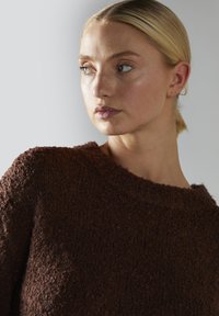 Młoda kobieta z blond włosami związanymi w kok, ubrana w teksturowany ciemnoszary sweter, patrzy w bok na jednolity jasny tle.