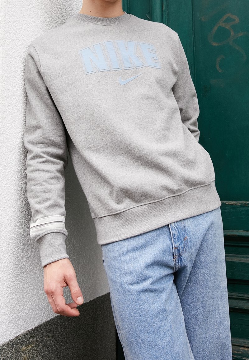 Personne portant un sweat-shirt Nike gris clair et un jean bleu clair, appuyée contre un mur blanc avec une porte verte en arrière-plan.