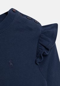 Prenda de punto azul marino con pequeño logo bordado, tres botones en el hombro y detalle de volante en la manga derecha.