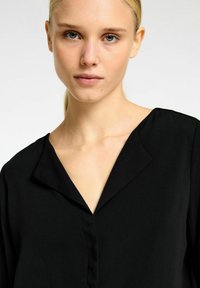 Sort bluse med V-hals, blødt stof og subtil glans. Har minimalistisk design med lange ærmer og en simpel knaplukning.