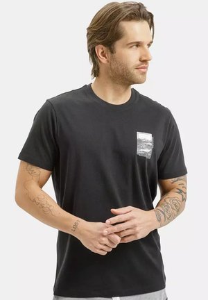 Mężczyzna o średniej długości brązowych włosach, ubrany w czarny t-shirt z małym białym nadrukiem na klatce piersiowej, pokazujący tatuaże na obu przedramionach, patrzący w prawo.