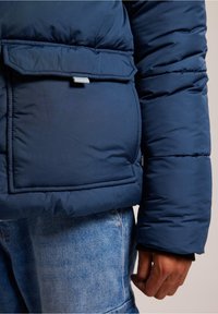 Marinblå pufferjacka med en stor framficka, slät textur, söm-detaljer och en åtsittande ärm. Denimjeans synliga nedan.