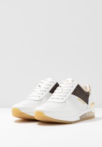 Scarpe da ginnastica bianche con tomaia in rete, accenti marroni e dettagli in oro. Presentano una suola spessa con una sezione trasparente per l'ammortizzazione.