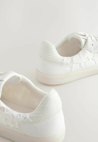 Hvide sneakers med en glat læderoverdel, blomsterlacemønster, struktureret hæl og gummisål. Klassisk snørebåndsdesign.