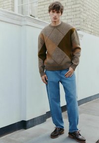 Maglione a fantasia marrone e verde, jeans azzurri chiari e scarpe scure con suola spessa; in piedi contro un muro chiaro.