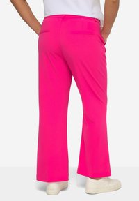 Pantalons évasés roses en tissu lisse, dotés de deux poches arrière, d'une taille et associés à des baskets blanches.