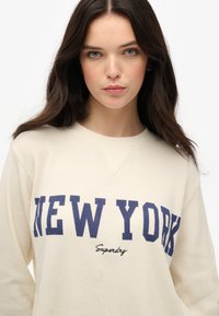 Felpa a costine crema con la scritta "NEW YORK" in lettere maiuscole blu navy e "Superdry" in corsivo sottostante. Collo rotondo e maniche lunghe.