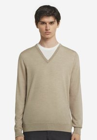 Beige v-ringad tröja med långa ärmar, tillverkad av mjukt material. Har en slät yta och ribbade mudd och nederkant. Lager på en vit skjorta.