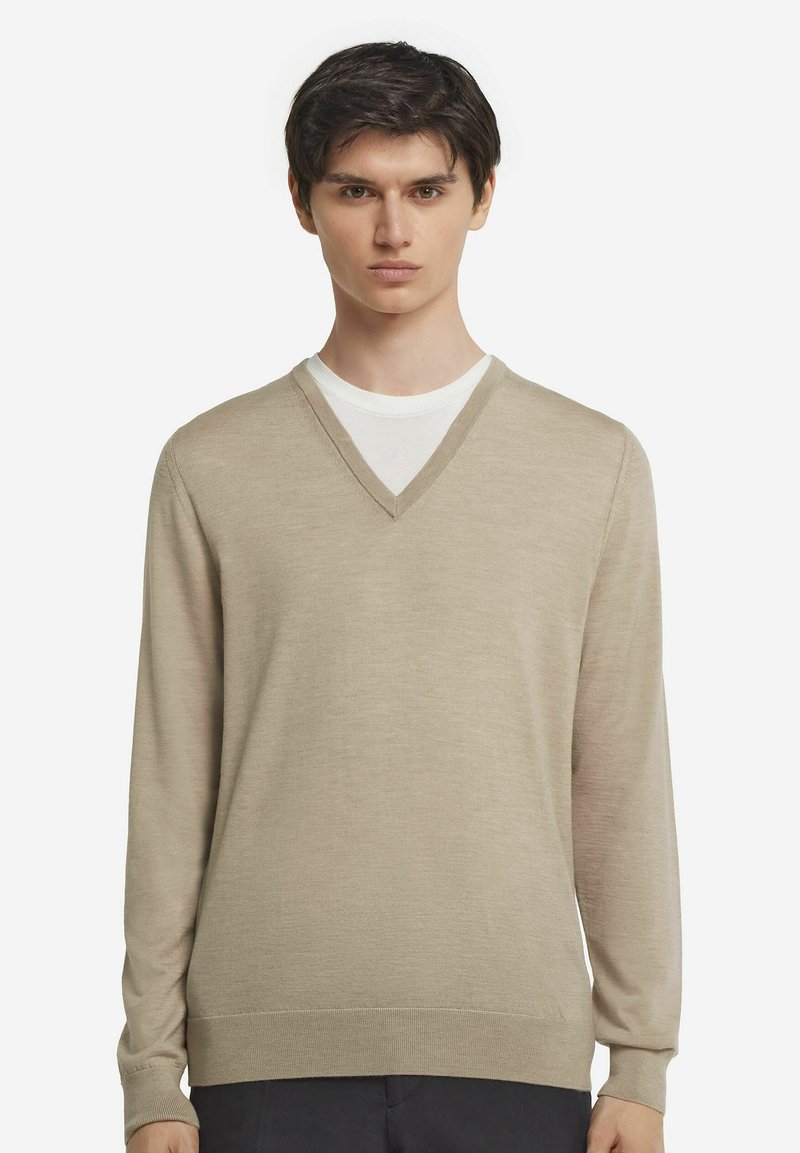 Beige v-ringad tröja med långa ärmar, tillverkad av mjukt material. Har en slät yta och ribbade mudd och nederkant. Lager på en vit skjorta.