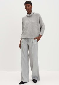 Grauer Wollrollkragenpullover, lockere Passform, kombiniert mit hellgrauen weiten Hosen. Schwarze Slippers mit dekorativen Ösen.