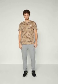 Camiseta marrón con estampado de camuflaje y mangas cortas, combinada con pantalones deportivos gris claro que presentan un logo. El modelo lleva zapatillas deportivas negras.